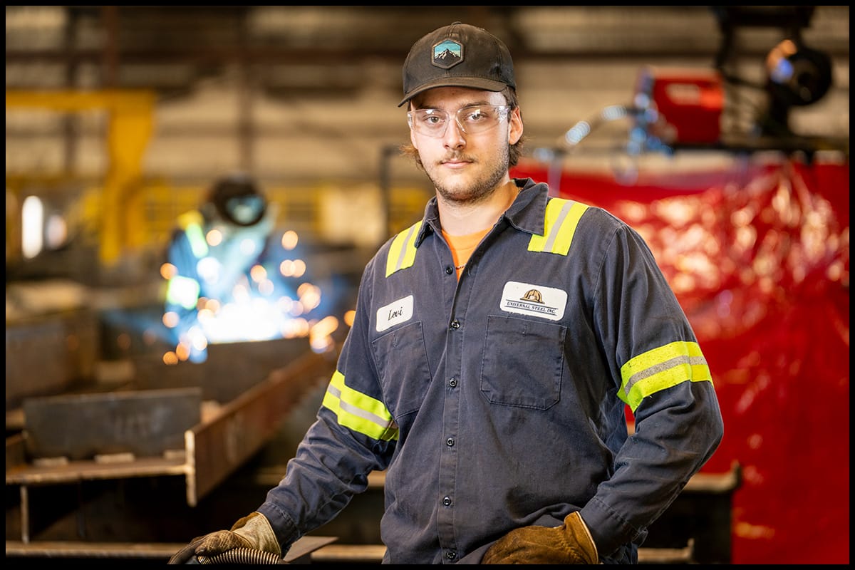 Portrait of an industrial worker inside an active fabrication environment. Industrial worker standing in a fabrication shop with welding activity in the background