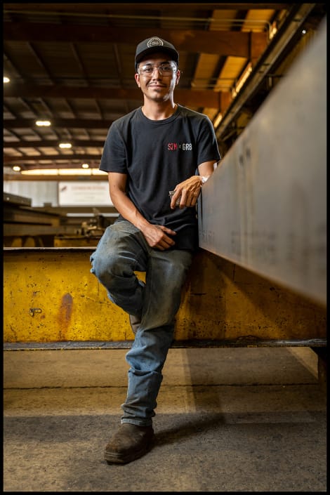 Environmental portrait captured inside the fabrication environment. Industrial worker seated beside structural steel inside a fabrication shop
