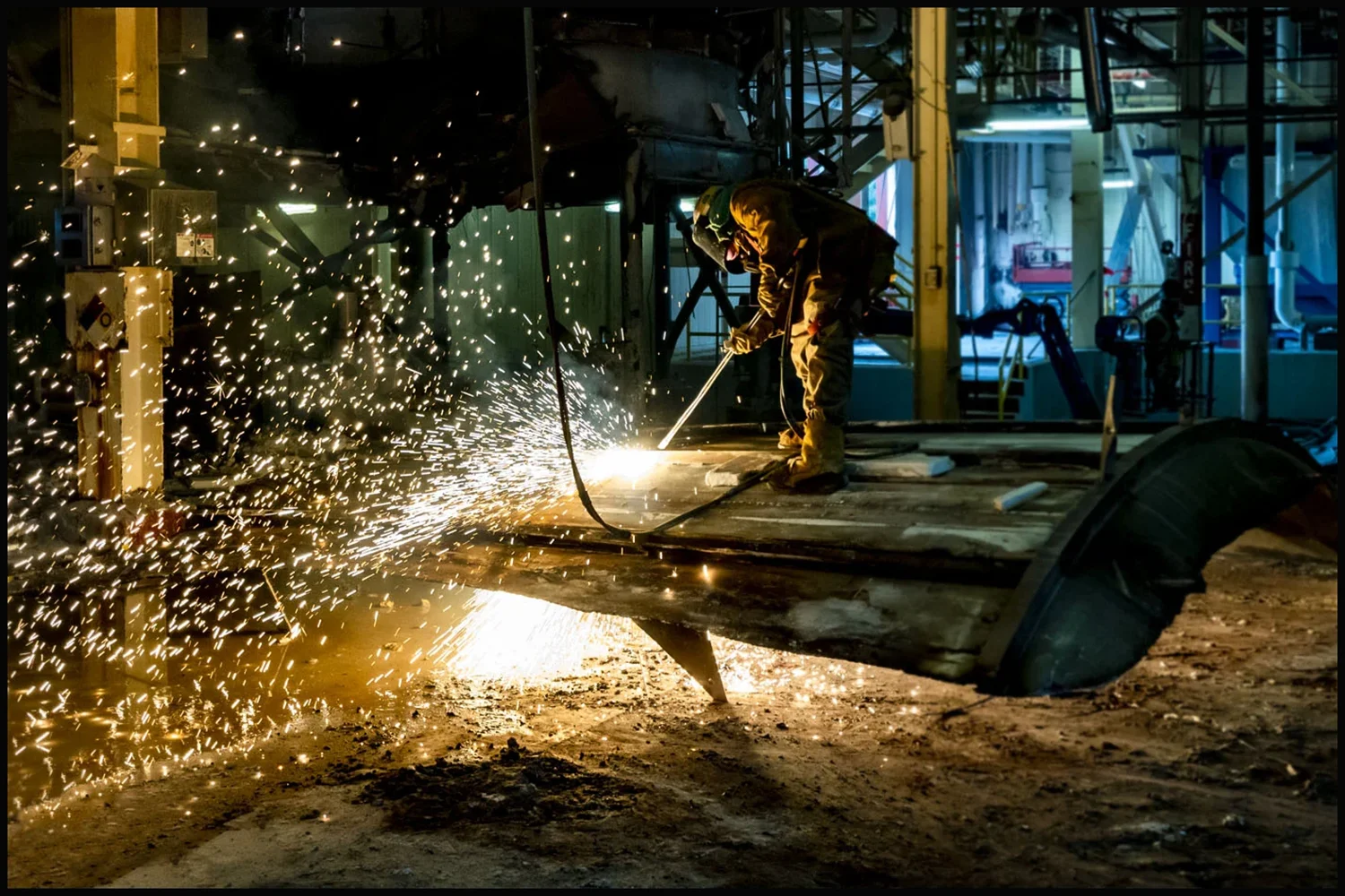 Industrial worker cutting steel with a torch as sparks fly across a manufacturing facility Industrial worker cutting steel with a torch as sparks fly across a manufacturing facility